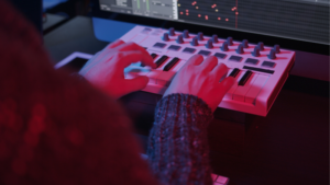 A person plays a compact MIDI keyboard with music production software visible on a computer screen in the background, possibly creating content for YouTube.