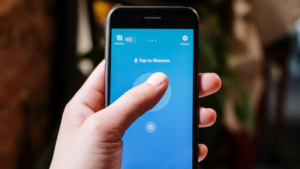 A person holds a smartphone displaying the Shazam app with the prominent "Tap to Shazam" button ready for music discovery.