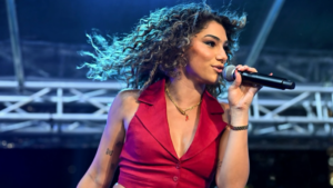 A singer with curly hair, wearing a sleeveless red top, holds a microphone and performs on an outdoor stage with metal trussing in the background.