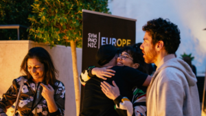 Four people interact outdoors; two are hugging, one is smiling and looking down, and another stands nearby. A sign behind them reads "SYMPHONIC EUROPE.