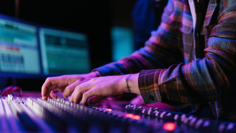 Person adjusting label-marked audio mixing console sliders, with computer screens in the background. The person is wearing a plaid shirt and has a tattoo on their wrist.