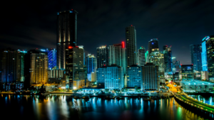 Nighttime cityscape showing illuminated high-rise buildings reflecting in the water, with busy streets buzzing as the Winter Music Conference brings energy to the scene under a cloudy sky.