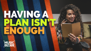 A woman with curly hair holds an open notebook and gestures, sitting on a couch. Text reads "Having a plan isn't enough. Music Biz 101." Colorful stripes are in the background, highlighting the importance of music marketing.
