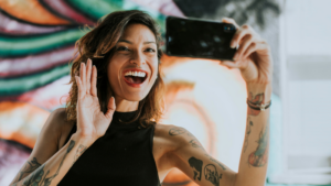 A smiling woman with tattoos waves at the camera while taking a selfie for her YouTube channel, capturing the moment in front of a colorful mural.