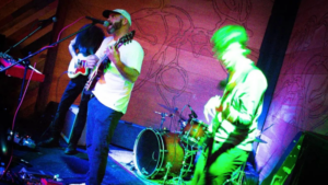 A band performs on stage with electric guitars, a keyboard, and a drum set under colorful stage lights, creating an intimate Tiny Desk vibe.