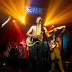 A band performs on a stage with colorful lighting at Bajo Circuito in Mexico City. One member sings and plays guitar shirtless, while others play instruments and sing.