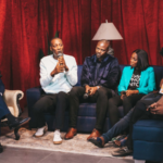 Five people sit on couches in front of red curtains; one person speaks into a microphone, while others listen or look at their phones. A lamp glows in the background, adding warmth to this Africa-inspired gathering.