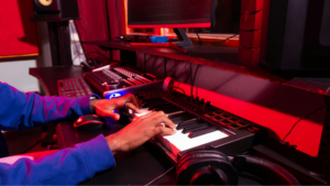 Person playing a digital keyboard in a music production studio, creating new tracks with samples on a computer, sound equipment, and headphones on the desk under red lighting.