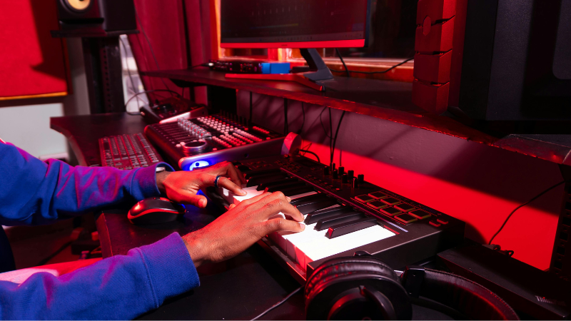 Person playing a digital keyboard in a music production studio, creating new tracks with samples on a computer, sound equipment, and headphones on the desk under red lighting.