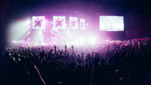 A large international crowd at a concert with hands raised, illuminated by stage lights and multiple screens displaying visuals.