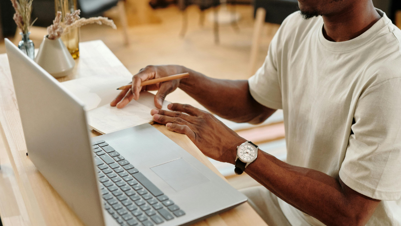 A contributor sits at a desk, writing on paper with a pencil next to an open laptop. The workspace includes a vase and various office items.