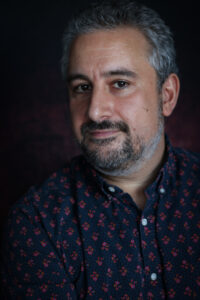 A middle-aged man with gray hair and a beard, wearing a patterned dark shirt, poses confidently against a dark, Ai-generated blurred background.
