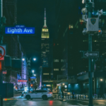 Night view of Eighth Avenue in New York City with street signs, a white car at an intersection, and the Empire State Building illuminated in the background—a vibrant scene near where A2IM hosts its annual music events.