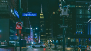 Night view of Eighth Avenue in New York City with street signs, a white car at an intersection, and the Empire State Building illuminated in the background—a vibrant scene near where A2IM hosts its annual music events.