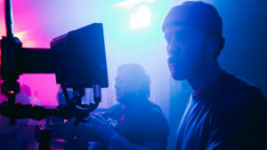 Two people work behind a video camera in a room with colorful blue and pink lighting, focusing on equipment and a monitor.