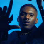 A man in dark clothing, reminiscent of many artists, holds his hands up in front of a blue background, with his face partially illuminated.