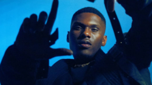 A man in dark clothing, reminiscent of many artists, holds his hands up in front of a blue background, with his face partially illuminated.