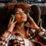 A person with curly hair sits in a chair, eyes closed, wearing large green headphones and a plaid shirt, appearing relaxed while listening to music in this editorial scene.