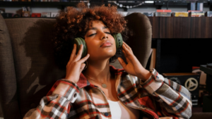 A person with curly hair sits in a chair, eyes closed, wearing large green headphones and a plaid shirt, appearing relaxed while listening to music in this editorial scene.