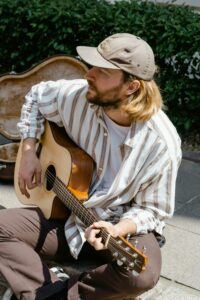 A man with long hair, wearing a cap and striped shirt, sits outdoors playing an acoustic guitar with an open guitar case behind him, dreaming of making it big in the music industry.