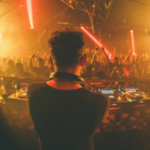 A DJ performs on stage with turntables in front, facing a crowded dance floor with raised hands and orange stage lighting—capturing the energy found at the best music venues for independent artists in Los Angeles.