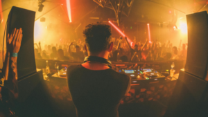 A DJ performs on stage with turntables in front, facing a crowded dance floor with raised hands and orange stage lighting—capturing the energy found at the best music venues for independent artists in Los Angeles.