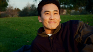 A person with short dark hair reclines on grass outdoors, smiling at the camera, with trees and greenery in the background.
