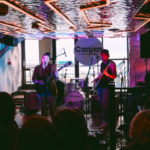 A live band performs onstage under neon lights at Canvas, with drums, guitars, and microphones visible; an audience watches in the foreground.