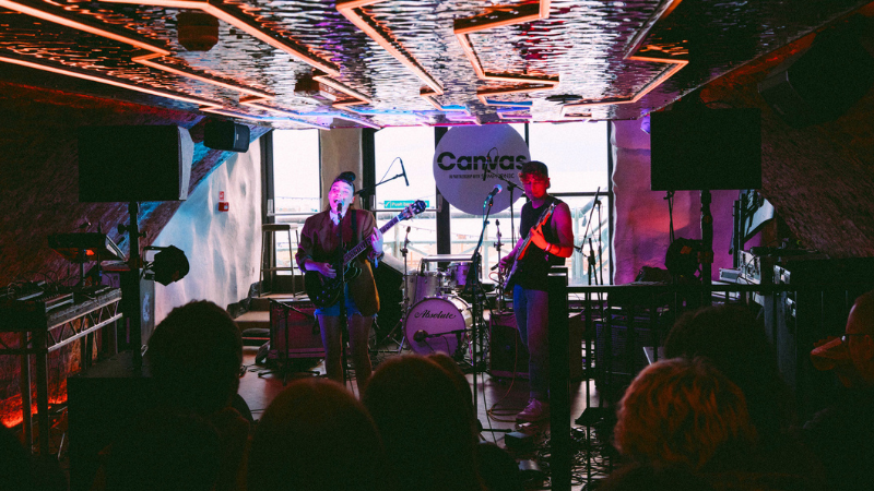 A live band performs onstage under neon lights at Canvas, with drums, guitars, and microphones visible; an audience watches in the foreground.