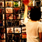 A person in a white shirt browses a rack filled with assorted postcards or greeting cards, thoughtfully selecting each one—as if searching for the perfect algorithm among the neatly arranged rows.