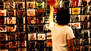 A person in a white shirt browses a rack filled with assorted postcards or greeting cards, thoughtfully selecting each one—as if searching for the perfect algorithm among the neatly arranged rows.