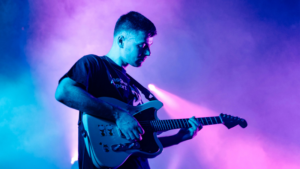 A man playing an electric guitar on stage with blue and purple stage lights in the background, captivating the crowd as part of his latest music release strategy.