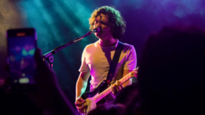 A person with curly hair, representing a record label brand, plays an electric guitar and sings into a microphone on stage under purple and yellow lights.