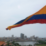 A large Colombian flag waves above a cityscape, where modern and historic buildings stand under a cloudy sky—an inspiring sight, especially during Circulart when creativity fills the air.