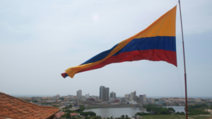 A large Colombian flag waves above a cityscape, where modern and historic buildings stand under a cloudy sky—an inspiring sight, especially during Circulart when creativity fills the air.