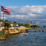 The Puerto Rican flag waves on a waterfront promenade lined with palm trees and buildings, as the calm sea and sky set the scene for celebrations like Premios Tu Música Urbano.