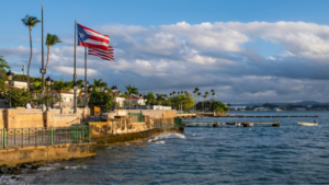 The Puerto Rican flag waves on a waterfront promenade lined with palm trees and buildings, as the calm sea and sky set the scene for celebrations like Premios Tu Música Urbano.