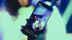 A person holds a smartphone displaying a live video stream of a guitarist performing on stage, with interface icons visible on the screen—showcasing the rise of music video platforms.