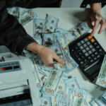 Person counting stacks of $100 bills with a calculator and money counter on a white table.