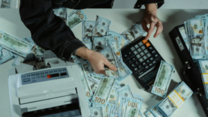 Person counting stacks of $100 bills with a calculator and money counter on a white table.