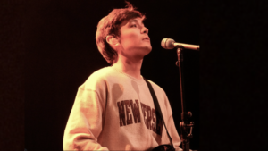 A person wearing a light-colored "NEW YORK" sweatshirt stands at a microphone onstage, looking upward under warm lighting, as if about to share a song from their playlist.