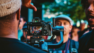 A camera on a stabilizer films a group of people outdoors, with several individuals visible in the background and someone adjusting the camera in the foreground—capturing content perfect for YouTube for musicians.