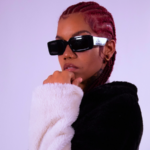 Black artists to watch: Person with braided hair, wearing dark sunglasses and a black-and-white fur jacket, poses against a plain light purple background with hand resting under their chin.