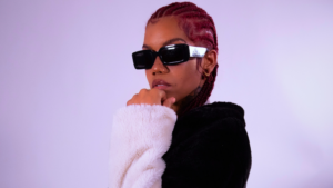 Black artists to watch: Person with braided hair, wearing dark sunglasses and a black-and-white fur jacket, poses against a plain light purple background with hand resting under their chin.