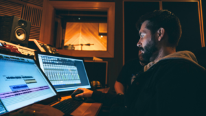 A man works at a computer in a recording studio, editing audio tracks on a screen with sound equipment and a recording booth visible in the background.