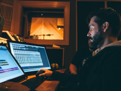 A man works at a computer in a recording studio, editing audio tracks on a screen with sound equipment and a recording booth visible in the background.
