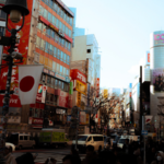 Busy city street in Japan with pedestrians, vehicles, and tall buildings featuring colorful billboards and advertisements for the Tokyo International Music Market. Japanese flags are visible on street lamps.