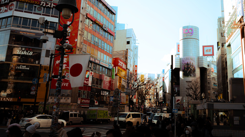 Busy city street in Japan with pedestrians, vehicles, and tall buildings featuring colorful billboards and advertisements for the Tokyo International Music Market. Japanese flags are visible on street lamps.