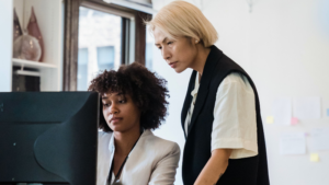 Two women in professional attire look at a computer monitor together in an office setting, one seated and one standing, collaborating on music industry mentorship programs.