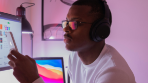 A person practicing mindfulness for musicians, wearing headphones and glasses, looks at a smartphone while sitting at a desk with computer screens in a softly lit room.